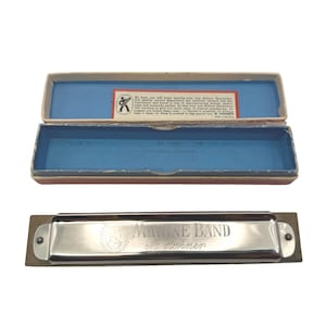 May include: A vintage Marine Band harmonica by M. Hohner, displayed with its original box. The harmonica has a silver finish and is set against a wooden base. The box is blue with a red and white trim and includes printed text.
