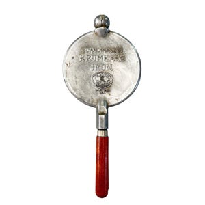 May include: A vintage Scandinavian Krumkake iron with a round, silver-toned cooking surface. The iron features the text "SCANDINAVIAN KRUMKAKE IRON" and a wooden handle. The handle is a rich, dark red color.