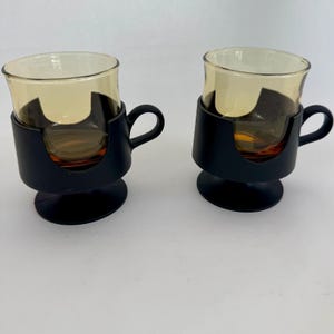 May include: Two glass cups with a light amber tint, each set into a matte black plastic holder with a circular base and a side handle. The glass inserts are removable from the black frames, featuring a smooth rim and a rounded body shape.