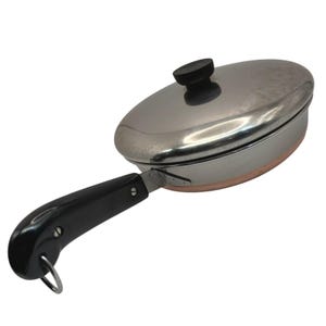 Puede incluir: Una sartén de acero inoxidable con base de color cobre y tapa a juego. La sartén cuenta con un mango negro con una anilla metálica para colgar en el extremo y un pomo negro en la tapa. La superficie exterior es pulida y muy brillante.