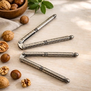 Puede incluir: Dos cascanueces plateados con mangos texturizados se muestran sobre una superficie de madera clara, junto con un cuenco de madera lleno de nueces. Los cascanueces están abiertos, mostrando su diseño.