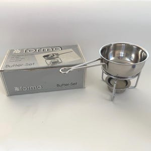 May include: A stainless steel butter warmer set with a small saucepan, stand, and box. The box is gray with the words "Butter-Set" and "forma" printed on it. The saucepan has a long handle and sits on a metal stand with a place for a tea light.