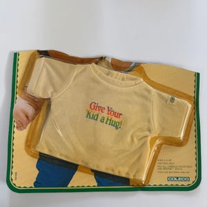 May include: A vintage Cabbage Patch Kids doll t-shirt in its original packaging. The cream-colored shirt has the text "Give Your Kid a Hug!" in rainbow colors. The packaging is green and yellow with the Coleco logo.