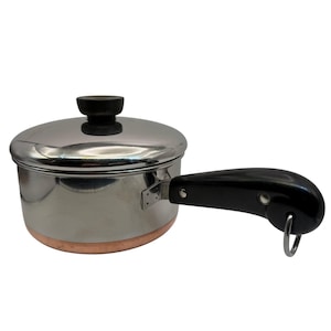 May include: Stainless steel saucepan with a copper bottom and a black handle. The pot has a lid with a black knob. The handle has a metal ring for hanging. The pot is designed for cooking.