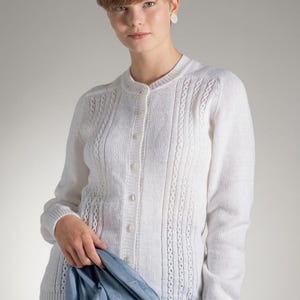 May include: A white long-sleeved button-down cardigan sweater featuring a vertical cable knit pattern and a ribbed crew neckline. The garment is crafted with a textured knit design and includes a row of matching white buttons down the front.