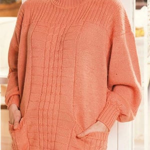 May include: A peach-colored long-sleeve knit sweater featuring a crew neckline, ribbed cuffs, and a ribbed hem. The sweater includes two front pockets and a vertical cable knit pattern running down the center of the torso.