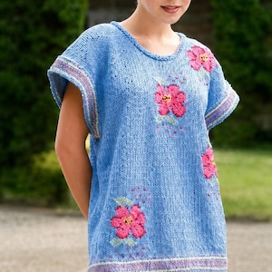 May include: A light blue knit short-sleeved top featuring a round neckline and embroidered pink floral motifs. The sleeves and hem are finished with thin, multi-coloured striped detailing. The garment is paired with plain white trousers.