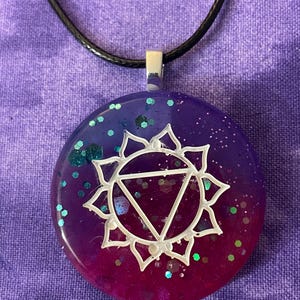 May include: A round pendant with a white chakra symbol on a gradient background of purple and magenta, with glitter accents. The pendant hangs from a black cord with a silver-colored bail.