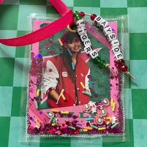 May include: A clear, plastic-covered photo featuring a person in a red jacket with white sleeves. The photo is decorated with pink glitter, colorful sprinkles, and the words "Tigers" and "Bayside" spelled out with letter beads. A pink ribbon is attached.