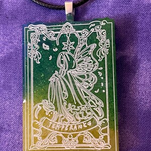 May include: A rectangular pendant with a black cord. The pendant features a white outline of a fairy-like figure with wings, a star, and the word "TEMPERANCE" on a banner. The background is a gradient of green and yellow.