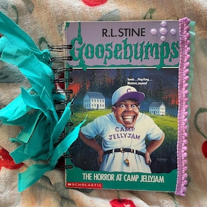 May include: A spiral-bound journal featuring the cover of "Goosebumps: The Horror at Camp Jellyjam" by R.L. Stine. The cover art depicts a cartoon character and two white houses. The journal is accented with teal ribbon and a pink trim.