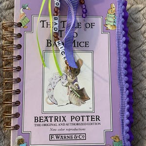 May include: A lavender spiral-bound notebook featuring the cover of "The Tale of Two Bad Mice" by Beatrix Potter. The cover art depicts a mouse with a broom. A beaded bookmark with the title of the book hangs from the top. The book has a purple pom-pom trim.