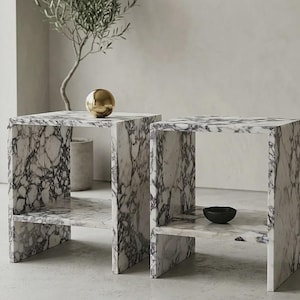 May include: Two marble side tables with a white and grey veined pattern. One table has a gold sphere on top, and the other has a small black bowl. The tables have a shelf and a square shape.