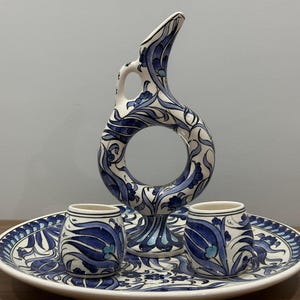 Könnte beinhalten: Ein Keramik-Servierset mit weißem Grund und blauen Blumenmustern. Das Set enthält einen hohen Krug mit offenem Zentrum, zwei kleine Tassen und ein rundes Tablett. Der Krug ist etwa 25 cm hoch.