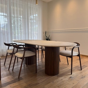 May include: An oval-shaped dining table with a light-colored top and a dark brown, ribbed base. Six wooden chairs with light-colored upholstered seats surround the table. The room has hardwood floors and sheer white curtains.