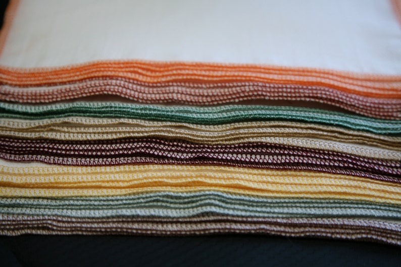 May include: A stack of folded fabric cloths in various colours, including orange, green, brown, and yellow. The cloths have a decorative stitched edge.