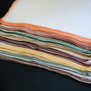 May include: A stack of colourful fabric squares in a variety of colours, including orange, green, brown, and yellow. The fabric squares are folded neatly and have a stitched edge.