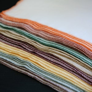 May include: A stack of colourful dishcloths in various shades of brown, green, yellow, and orange. The cloths are folded and layered, creating a textured and visually appealing display.