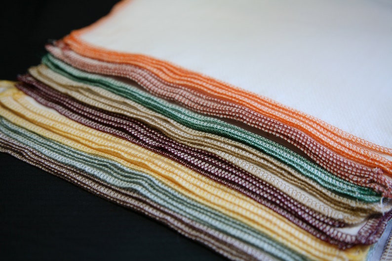 May include: A stack of colourful dishcloths in various shades of brown, green, yellow, and orange. The cloths are folded and layered, creating a textured and visually appealing display.