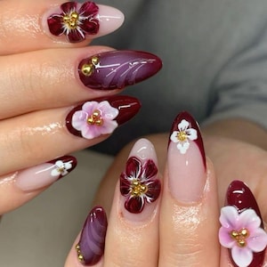 May include: Close-up of a hand with several nails painted in a deep burgundy color. Each nail is adorned with a 3D flower, some with white petals and others with pink petals. The flowers have gold accents.