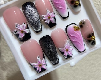 May include: A set of ten artificial nails in a white tray. The nails feature various designs, including pink and black glitter, floral accents, and abstract patterns. Some nails have gold embellishments.