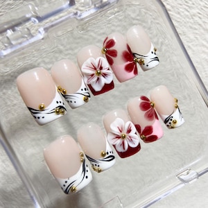 May include: A set of artificial nails in a clear plastic case. The nails feature a light pink base with white tips, black and gold accents, and 3D flower designs. Some nails have red floral patterns.