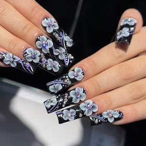 May include: Long, square-shaped black acrylic nails with 3D white and gray flowers, purple accents, and rhinestones. The nails are decorated with a floral design and are a fashionable accessory.