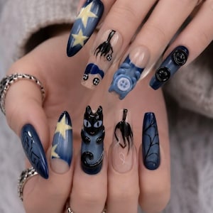 Pode incluir: Unhas postiças longas em formato amêndoa e bailarina com base azul-escura e vários designs, incluindo estrelas amarelas, um gato preto, teias de aranha, olhos de botão e um rosto de personagem. O conjunto inclui elementos 3D como botões e um gato.