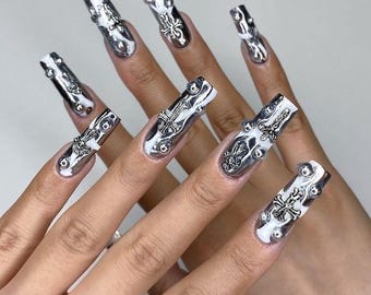 Pode incluir: Unhas longas, prateadas e brancas, com adornos metálicos ornamentados. As unhas têm formato de caixão, com base branca e detalhes prateados. O design inclui detalhes de cruzes e tachas.