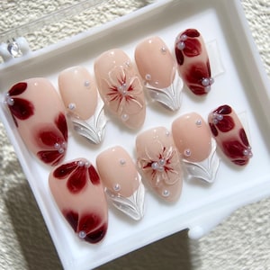 May include: A set of artificial nails in a clear plastic case. The nails feature a nude base with burgundy floral designs and pearl accents. Some nails have a 3D flower design. Silver leaf-shaped accents are also present.