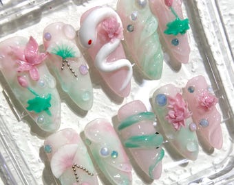 Può includere: Un set di dieci unghie artificiali rosa e verdi con disegni floreali e di serpente in 3D. Le unghie sono decorate con strass e glitter.
