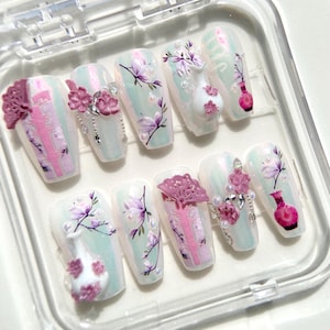 May include: A set of ten white press-on nails with a pastel blue and pink floral design. The nails feature 3D embellishments including flowers, vases, and fans.