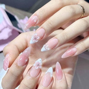 May include: A close-up of a set of long, almond-shaped nails. The nails are painted with a gradient of pink and white, with a 3D flower and rhinestone accents. The nails have a glossy finish.