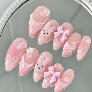 Könnte beinhalten: Rosa und weiße Nagelkunst mit einem Hasen- und Blumendesign. Die Nägel sind lang und oval geformt.