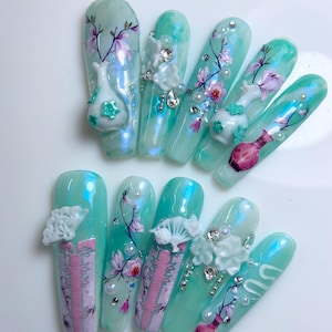 May include: A set of ten long, oval-shaped artificial nails. Each nail is painted with a light teal iridescent polish and decorated with floral designs, 3D embellishments, and rhinestones. Some nails feature small vase-shaped ornaments.
