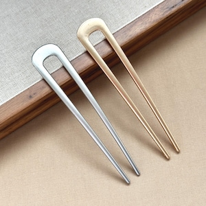May include: Two metal hair sticks. One is silver, the other is gold. Each has a U-shaped top and a long, slender pin. They are resting on a light beige surface and a wooden tray.
