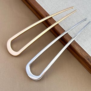May include: Two metal hair pins, one gold and one silver. Both have a U-shaped design with long, straight prongs. The pins are displayed on a neutral surface with a wooden accent. The pins are likely for styling hair.