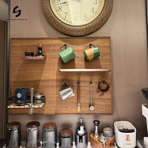 May include: A wooden pegboard with a variety of items hanging on it, including mugs, a coffee grinder, a whisk, and a scoop. There are also several jars of coffee beans and a coffee maker on the counter below the pegboard.