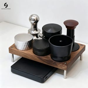 Op de afbeelding: Een houten standaard met vier zilveren poten bevat een verzameling espresso-accessoires, waaronder een zilveren tamper, een zilveren doseerring, een zwarte distributeur, een zwarte doseertrechter en een zwarte tamper met een donkerhouten handvat.