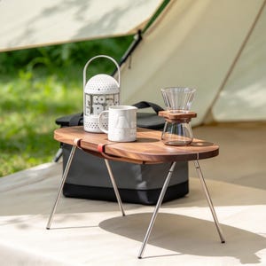 May include: A portable wooden folding table with four thin metal legs, featuring a white lantern, a white ceramic mug, and a glass coffee dripper set on top. The table has a smooth, oval-shaped walnut surface with a small red strap detail.