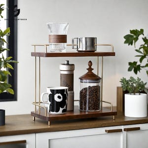 May include: Two-tiered wooden shelf unit with a gold metal frame. The top shelf has a glass carafe, a silver mug, and a wooden lid. The bottom shelf has a black and white mug, a silver grinder, and a glass jar filled with coffee beans.