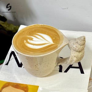 Puede incluir: Una taza de café de cerámica moteada llena de un café con leche, con un diseño de hoja en la espuma. El asa tiene una forma texturizada única. La taza está sobre una superficie blanca con el texto "SQUIRRELARTDECO".