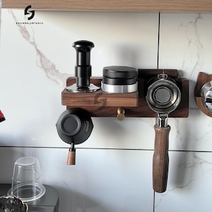 Puede incluir: Una estación de espresso de madera con un tamper, un portafiltro y otros accesorios de café. La estación está montada en una pared de baldosas de mármol blanco. Los accesorios son negros y plateados.