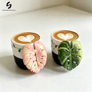 Pode incluir: Duas chávenas de café em cerâmica com latte art em forma de coração, cada uma com um adorno de folha de monstera. Uma folha é cor-de-rosa claro com pintas amarelas, a outra é verde-escura com variações mais claras e pontos brancos.