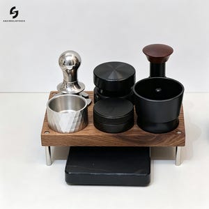 Op de afbeelding: Een houten koffiestation-organizer met vier metalen poten, met een zilveren metalen tamper, een zilveren gefacetteerde doseerbeker, twee zwarte ronde koffieverdelers en een zwarte trechter met een donkerhouten handvat boven een zwarte weegschaal.