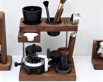 May include: A dark wood espresso station organizer featuring a two-tier design with slots for a portafilter, a tamper, cleaning brushes, and a distribution tool. The stand includes a black cup, a circular holder for filters, and metal support rods.
