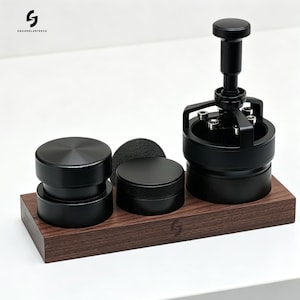 May include: A coffee distribution and tamping set featuring three matte black metal tools on a rectangular dark wood base. The set includes a cylindrical tamper, a circular distributor, and a spring-loaded levelling tool with visible silver bolts.