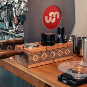 May include: Wooden espresso station with a portafilter, tampers, and a metal holder. The station has a decorative pattern and the text "EQUIPMENT & CO". A coffee machine is in the background.