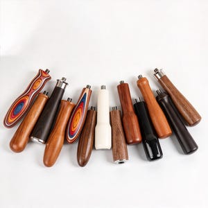 May include: A collection of twelve cylindrical handles arranged in a semi-circle. The handles are crafted from various materials including solid wood in light and dark brown tones, black polished material, cream-colored material, and multi-colored laminated wood with vibrant concentric patterns. Each handle features a small, threaded metal stud at the top for attachment.