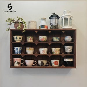 Peut inclure: Une étagère en bois brun foncé avec 18 compartiments, chacun contenant une petite tasse ou un bol en céramique. Les tasses et les bols sont décorés de motifs et de designs variés, notamment des fleurs, des animaux et des formes géométriques. L'étagère est fixée à un mur blanc.